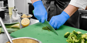 Blue-gloved hands chop green herbs on a green cutting board in a professional kitchen.